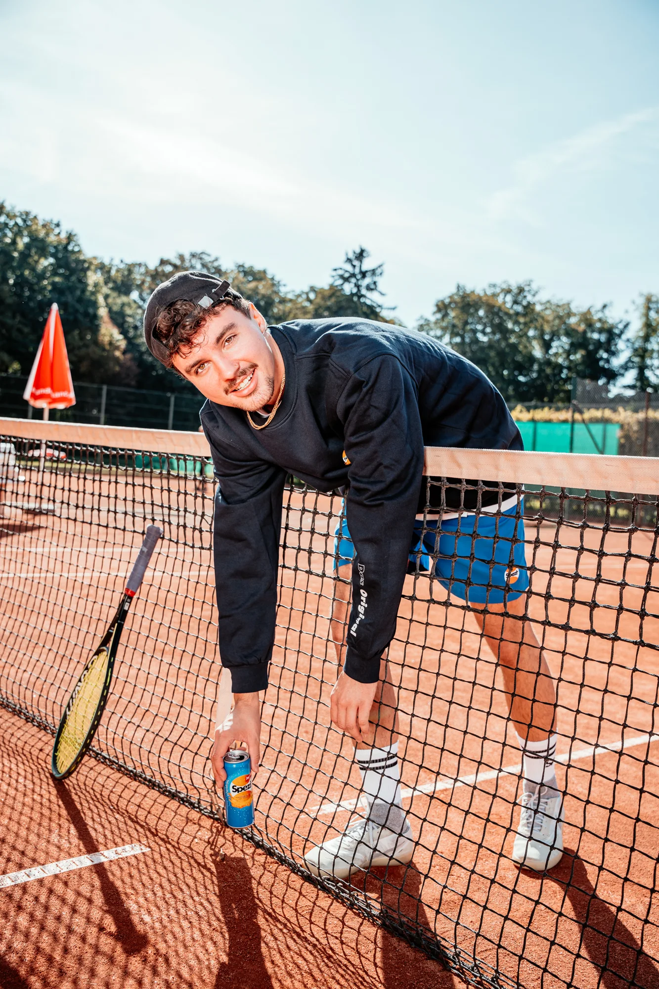 Lifestyle-Motiv mit Spezi-Dose und Marken-Pulli am Tennisnetz