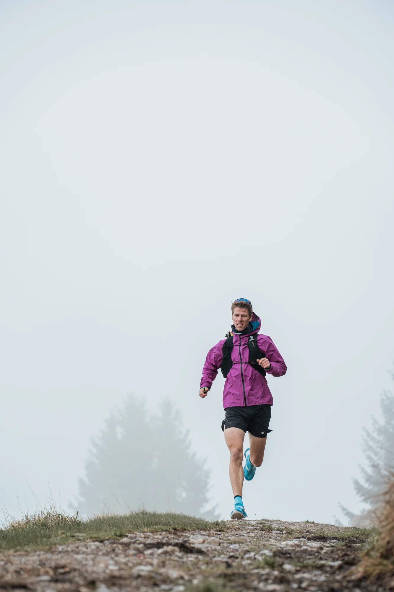 Trailrunner in violetter Jacke auf einem felsigen Weg im Bergnebel